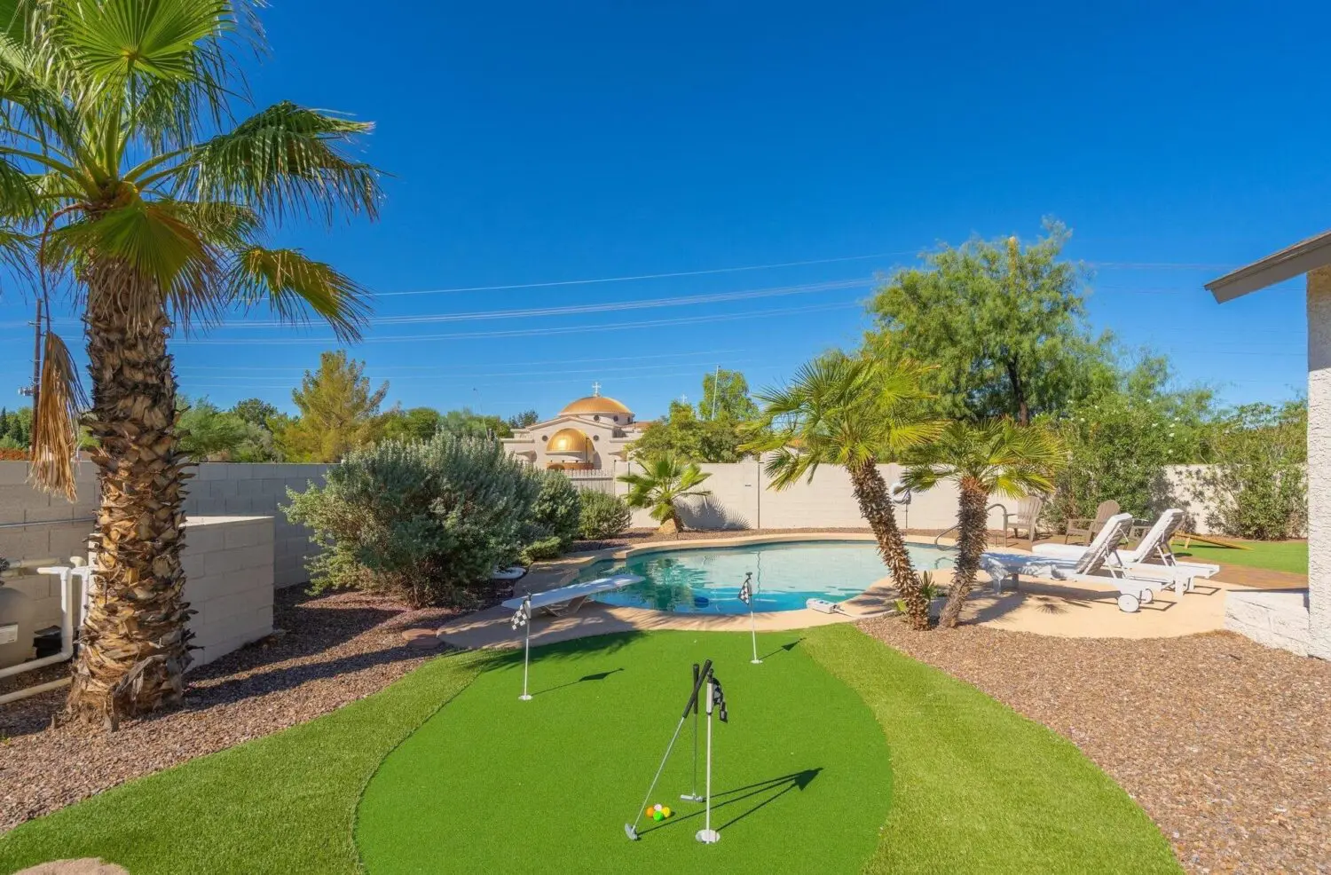 A backyard in El Mirage with palm trees, a small putting green made of artificial grass, and a swimming pool surrounded by lounge chairs under a clear blue sky.