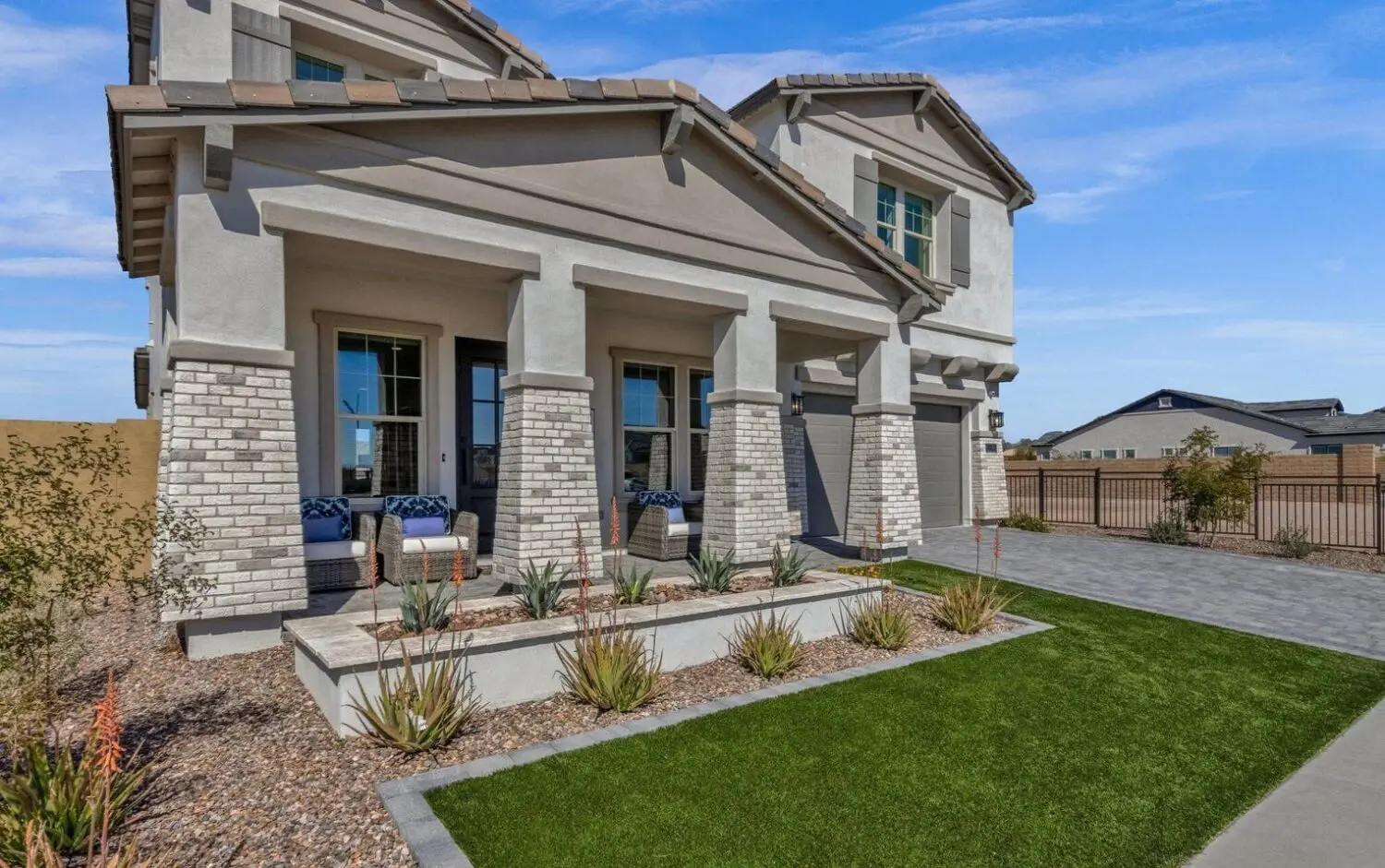 Modern two-story house in Litchfield Park with gray and white exterior, brick accents, three front porch columns, blue cushioned chairs, manicured lawn featuring artificial turf and desert landscaping, plus a paved driveway under a blue sky.