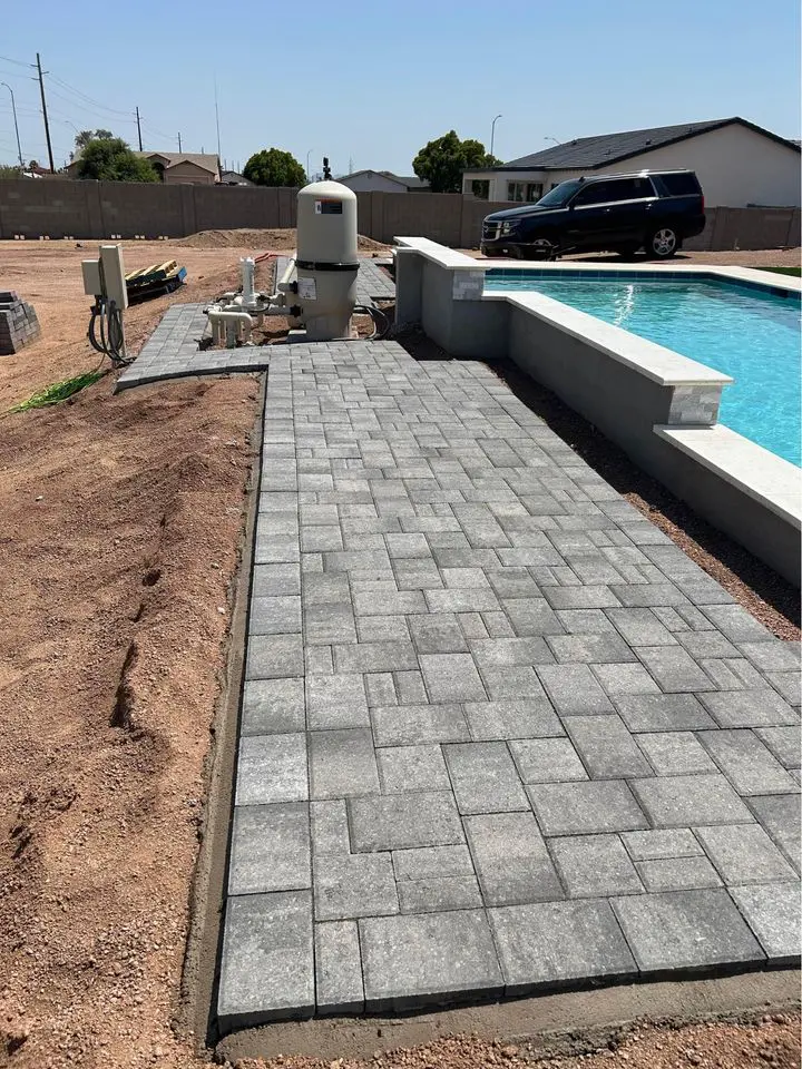 A newly installed gray stone walkway, showcasing expert paver installations, leads to a bright blue swimming pool. Construction equipment is nearby, and a black SUV is parked in the background. The area, typical of Buckeye AZ, is surrounded by a dry landscape and some residential houses.