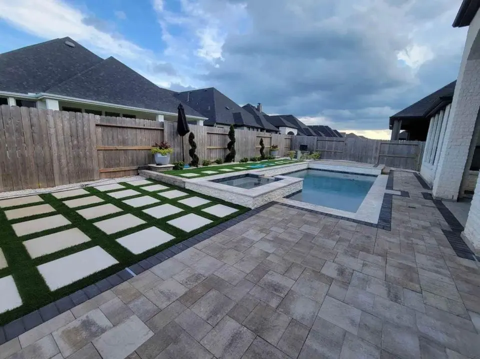 Modern backyard in Buckeye featuring a small pool and hot tub, surrounded by stone and brick paver paths. A wooden fence encloses the area adorned with decorative sculptures and potted plants. An overcast sky looms above suburban houses in the background, showcasing expert paver installations fit for AZ terrain.