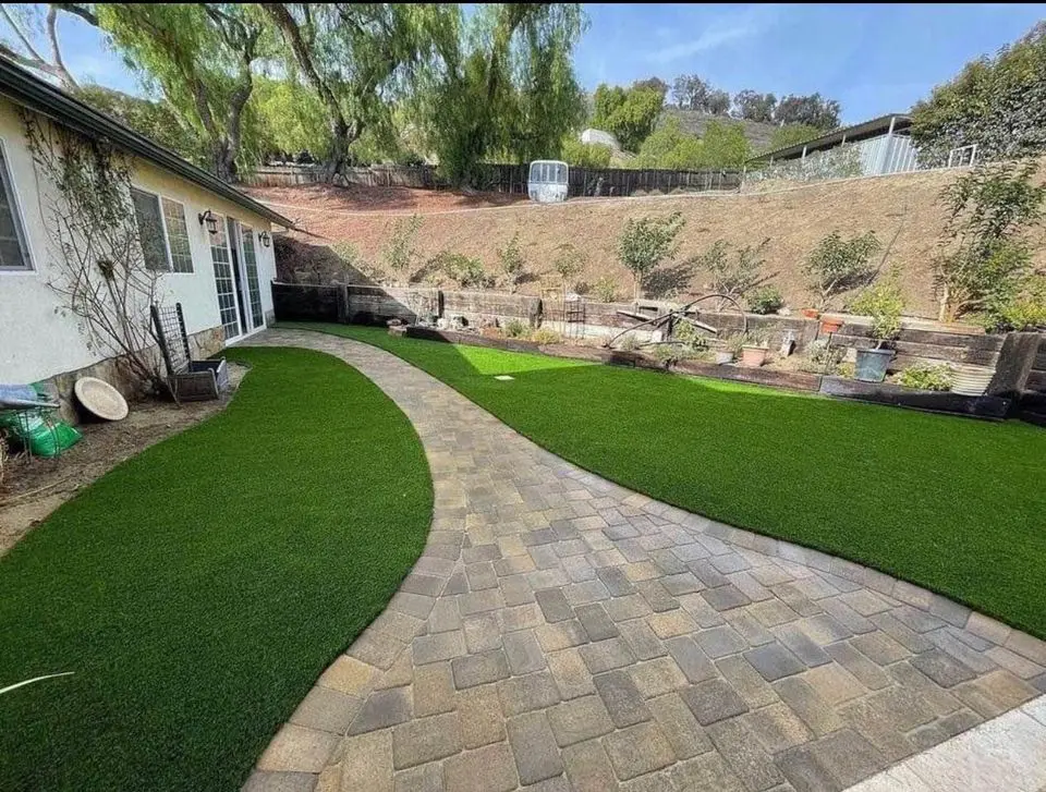 A backyard in Buckeye, AZ boasts a stone pathway leading to a house on the left. The area features artificial grass, lush plants, and a hillside in the background. Several potted plants and vibrant garden beds line the edges, showcasing beautiful stone and brick pavers.