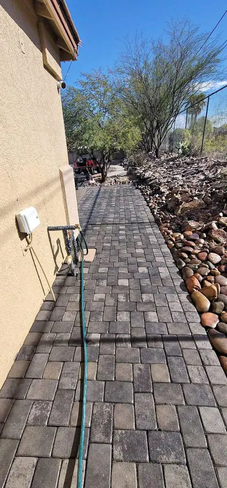 A paved walkway made of square stones, using stone and brick pavers, runs alongside a tan building. The path is bordered by a rock garden and shrubbery, seamlessly enhancing the outdoor space. A green garden hose is laid across the path under a clear blue sky.