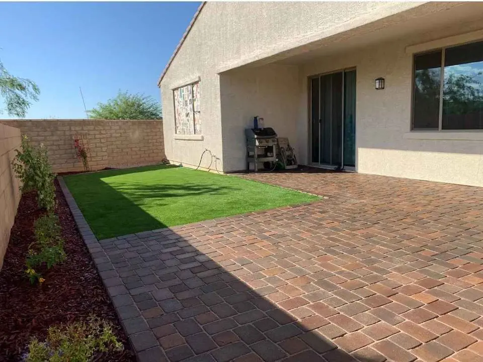 A backyard in Buckeye, AZ showcases a brick patio with paver installations and a small grassy area. The light-colored single-story house has a sliding glass door and window. A barbecue grill rests against the house wall, surrounded by plants along the border.