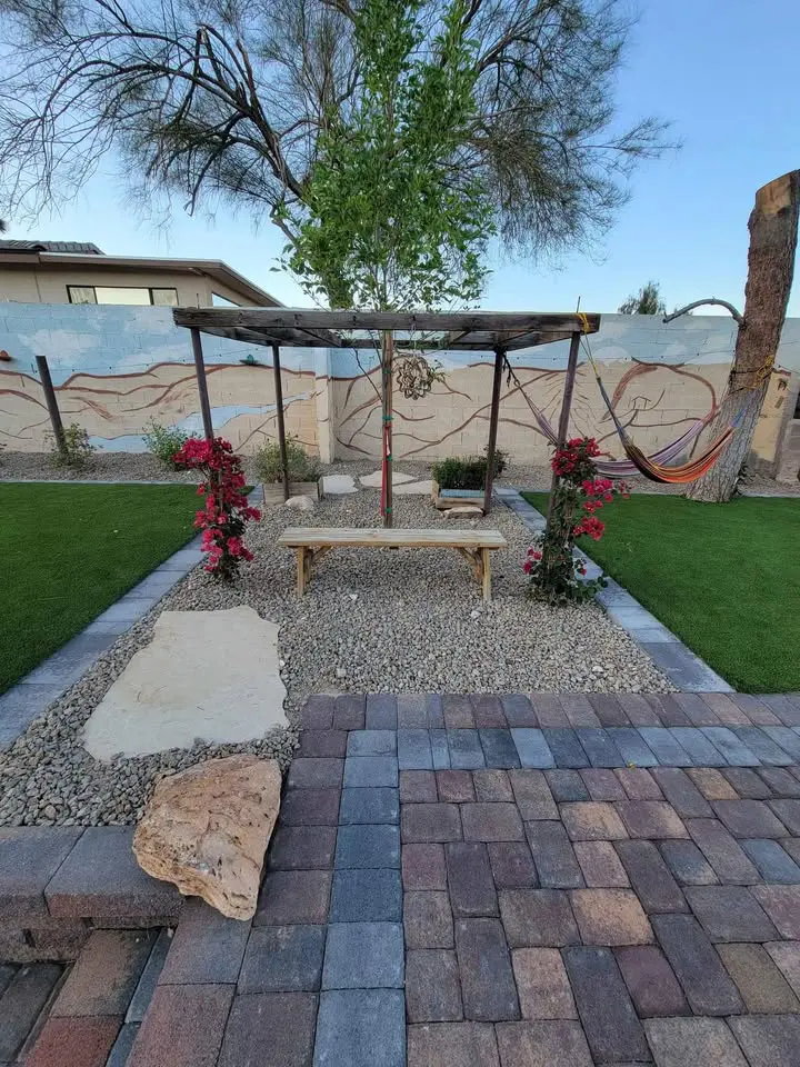A backyard garden in El Mirage features a pergola with flowering vines, a wooden bench on gravel, and a hammock to the right. Green artificial grass borders stone paths and a mural-painted fence with trees visible in the background.