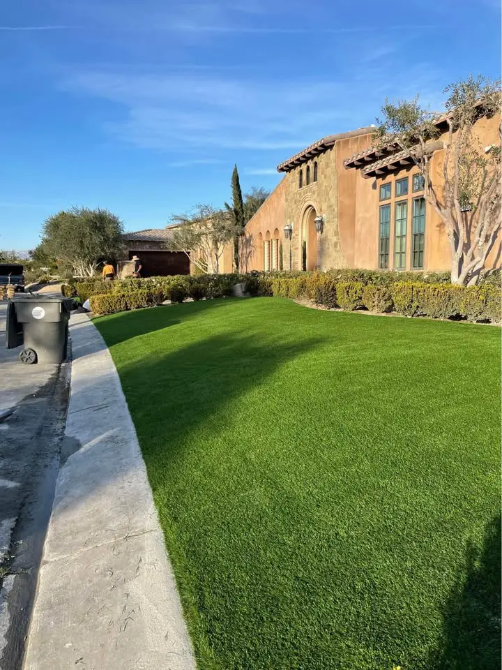 A well-manicured green lawn, created with artificial turf Peoria AZ, stretches in front of a large Mediterranean-style house with tan stucco walls, arched windows, and trimmed bushes along the sidewalk on a sunny day.