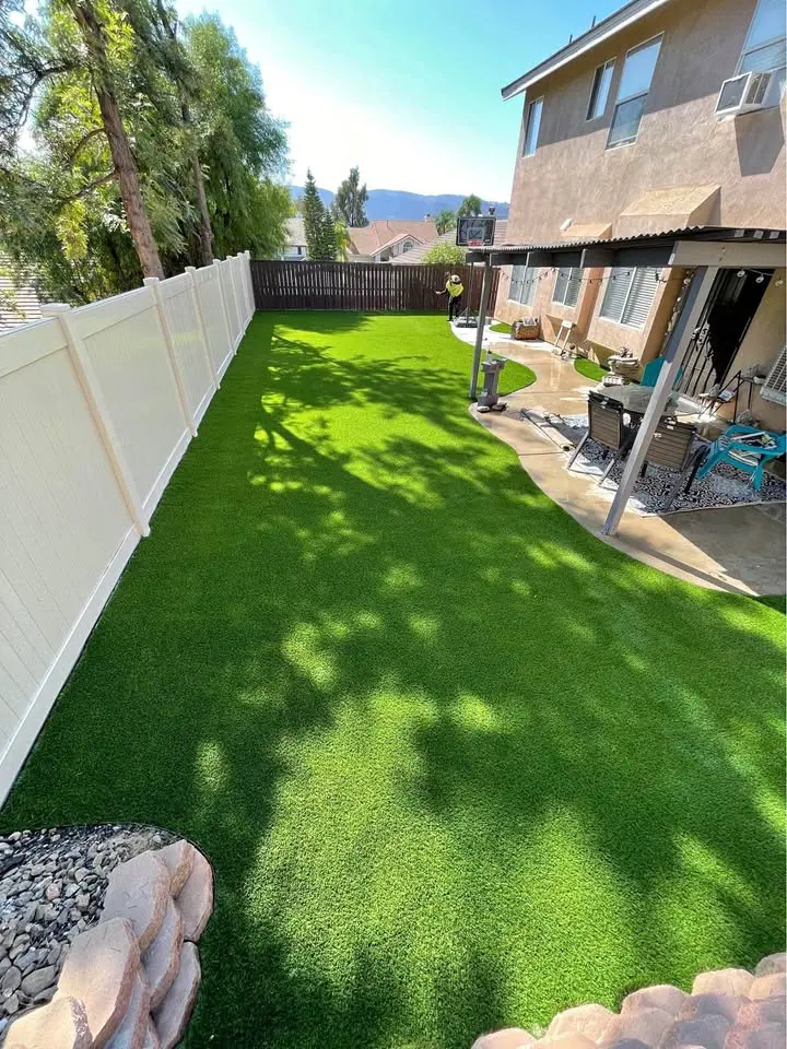 A backyard with bright green artificial turf from professional artificial grass installations, a white fence along the left side, patio furniture under a covered area, and trees casting shadows beside a beige house on a sunny day.