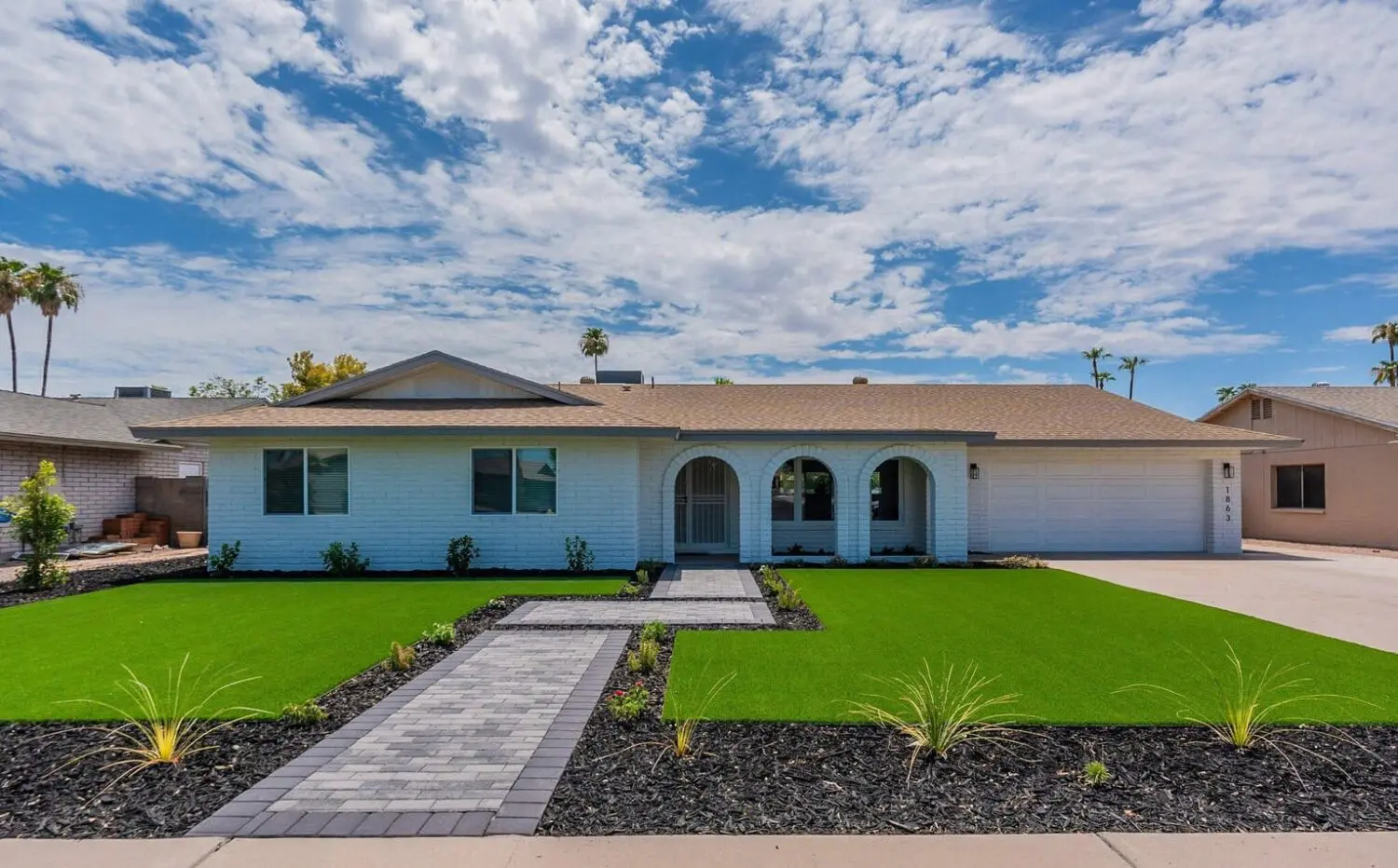 Single-story white house with a tan roof, three front arches, and a two-car garage in Sun City AZ. The yard features artificial grass installation with a brick walkway, small shrubs, and a clear blue sky with scattered clouds above.