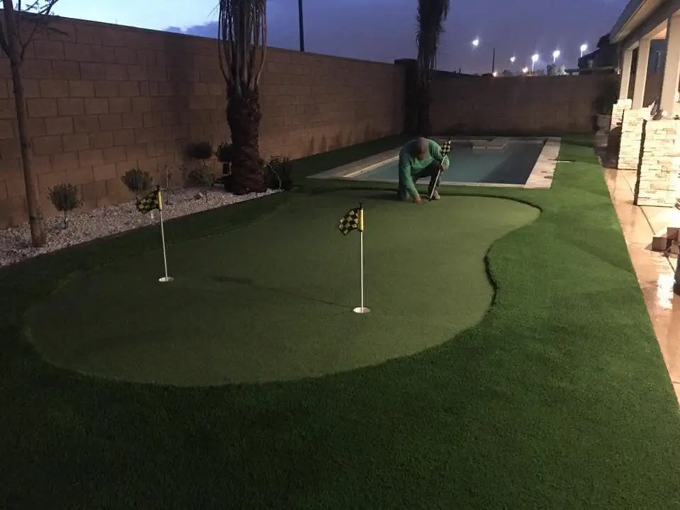 A person is installing a putting green made of synthetic turf with two golf holes and flags in a backyard next to a long, narrow swimming pool at dusk.