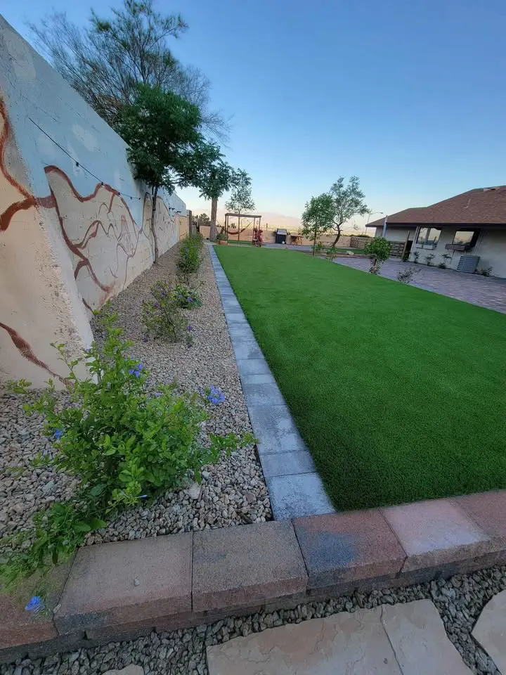 A neatly landscaped backyard with synthetic turf bordered by bricks, decorative gravel, and garden plants along a painted wall. Trees and a playground set are visible in the background near a house.