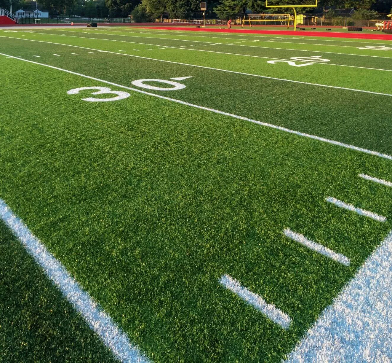 A close-up view of an American football field in Buckeye, Arizona, showing the 30-yard line with vibrant artificial grass installations and white painted yard markers. In the background are the goalposts and a surrounding track, with trees and stands visible further back. Get a free service quote today.