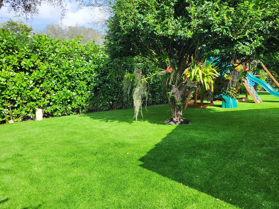 A sunny backyard in Litchfield Park with lush artificial turf, tall hedges, and a tree with hanging moss. In the background, a wooden playset with a slide and swings is partly visible under the shade.