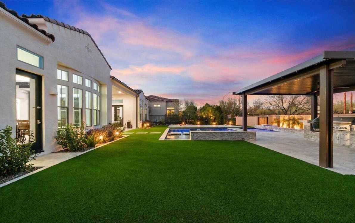 Modern El Mirage backyard at sunset with artificial grass, a swimming pool, a covered patio and outdoor kitchen, plus soft landscape lighting along the side of a white house. Trees and shrubs border the yard, highlighting expert artificial turf installation.