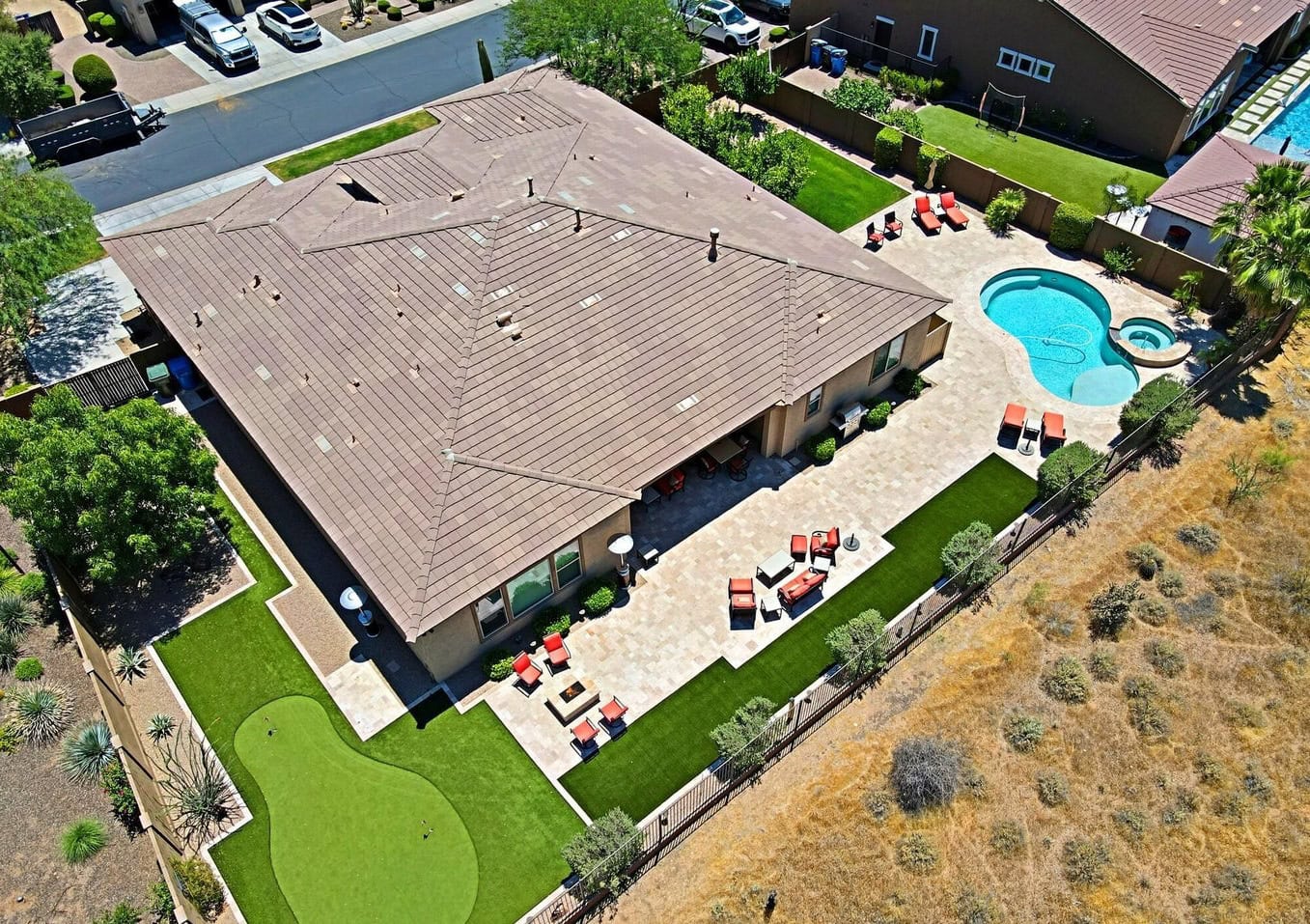 Aerial view of a modern house in Litchfield Park with a tiled roof, spacious patio, outdoor seating, putting green featuring artificial turf, and a swimming pool in a landscaped backyard bordered by dry, grassy areas.