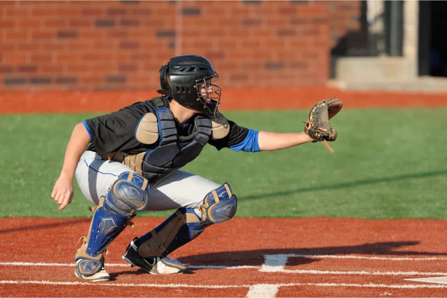 A baseball catcher in full gear crouches behind home plate on a sunny day, ready to catch a pitch, with vibrant synthetic turf beneath him for a flawless playing surface.
